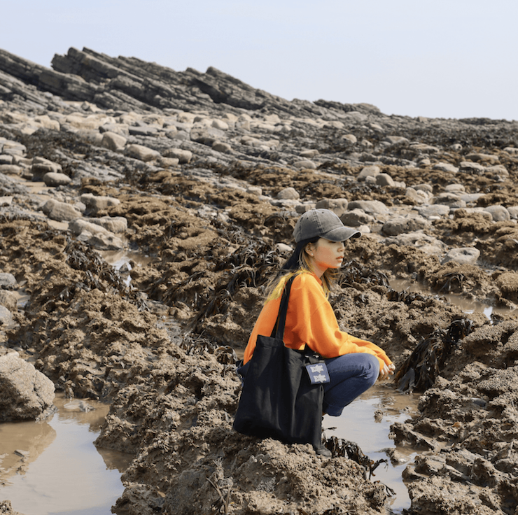 Angela in an orange jumper crouched at a rocky seaside. She wears a black tote bad and a grey cap.