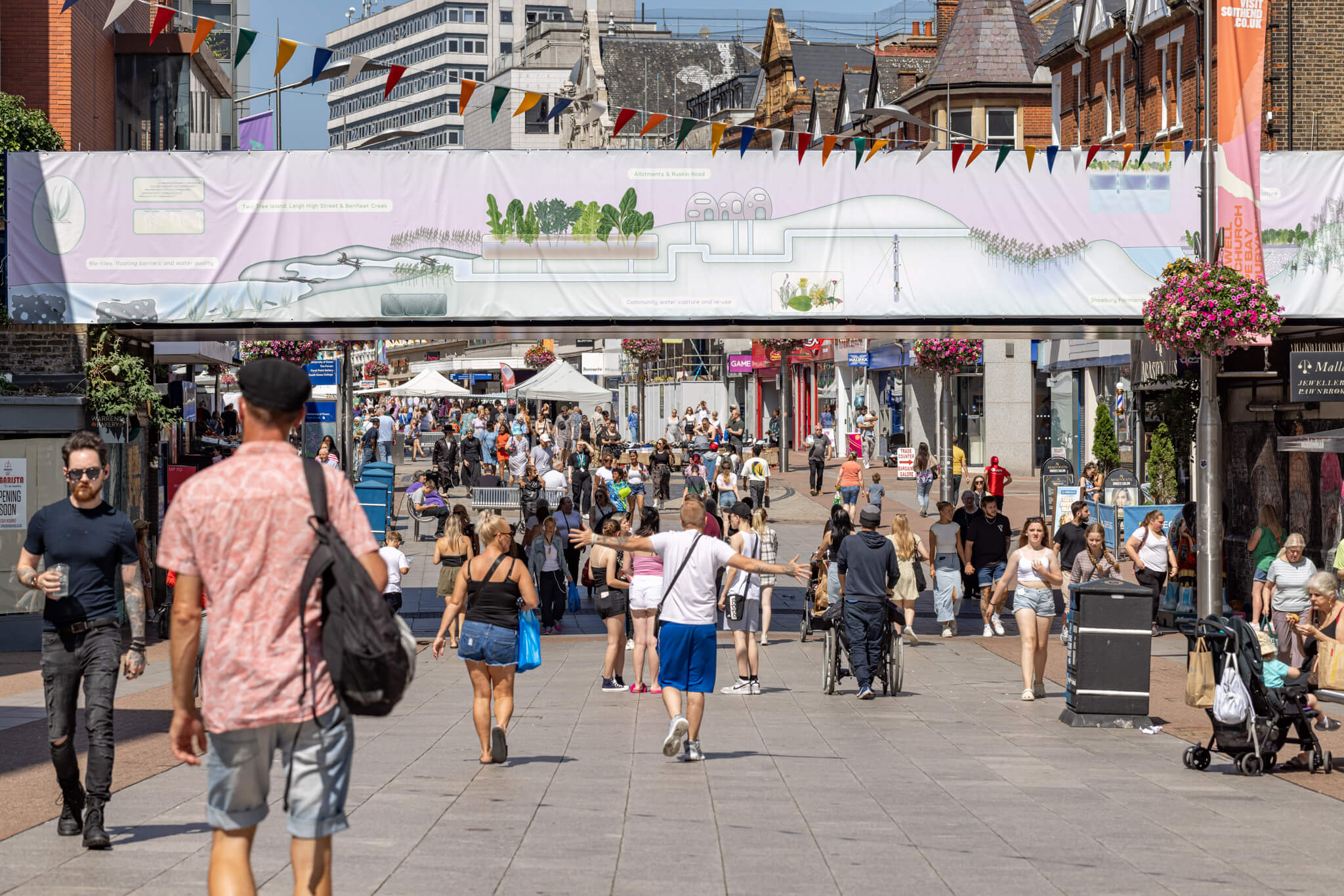 a view of a busy seaside high street with people walking. Above them is a railway bridge, decorated with a horizontal banner by the artist. It depicts a pastel green and pink landscape of a coastal town and illustrates various nature-based ecologial solutions to flood risks and coastal erosion, such as water pipes, vegatation, weather stations