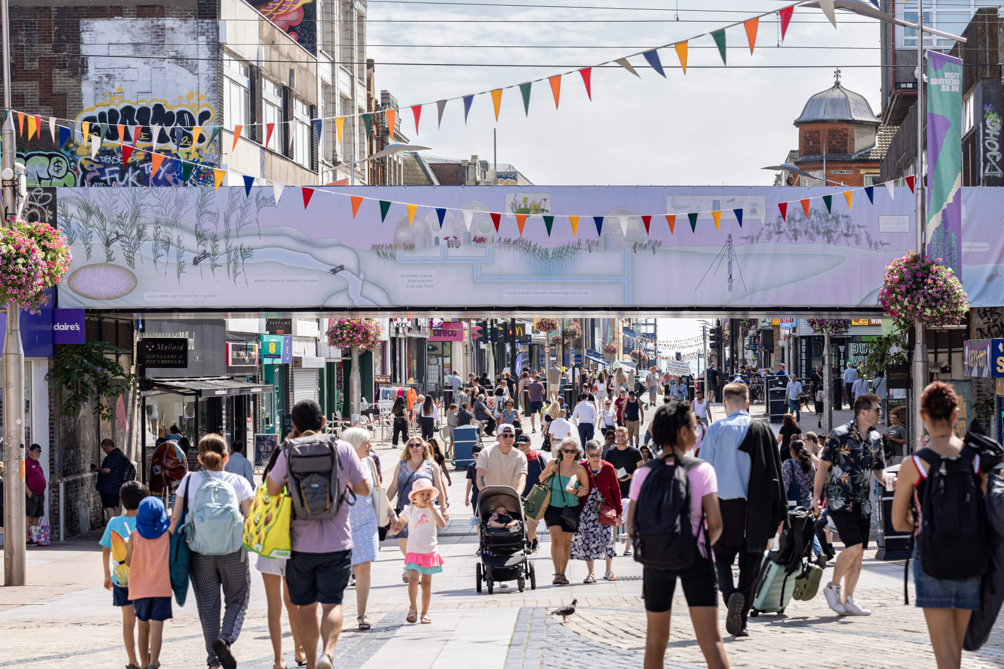 from the other direction of the road, a view of a busy seaside high street with people walking. Above them is a railway bridge, decorated with a horizontal banner by the artist. It depicts a pastel green and pink landscape of a coastal town and illustrates various nature-based ecologial solutions to flood risks and coastal erosion, such as water pipes, vegatation, weather stations